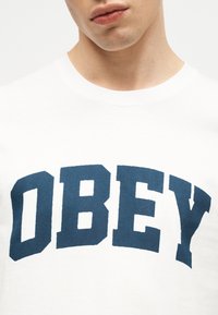 Κοντινό πλάνο ενός ατόμου που φοράει λευκό πουκάμισο με μεγάλα, έντονα μπλε γράμματα που σχηματίζουν τη λέξη «OBEY» στο στήθος.