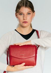 Bolso bandolera de cuero rojo con acabado texturizado, cierre magnético en la solapa y correa ajustable. Logotipo dorado en la parte frontal.