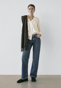 Kremowa koszula zapinana na guziki, ciemnobrązowy szalik zarzucony na ramię, niebieskie jeansy szerokimi nogawkami, czarne płaskie buty z frędzlami, neutralne tło.