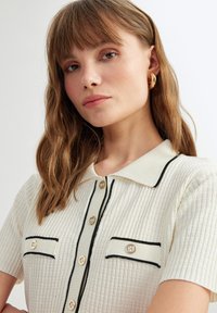 Polo shirt a coste crema con maniche corte, bordo nero lungo il colletto e le tasche, e bottoni decorativi.