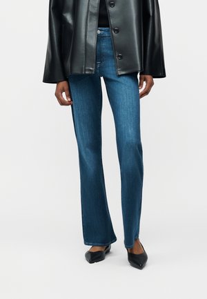 Personne portant un jean droit bleu, des ballerines noires pointues et une veste en cuir noire boutonnée, debout devant un fond uni.