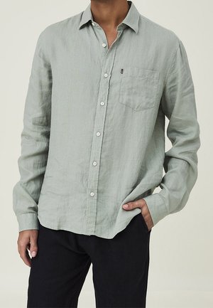 Homme portant une chemise gris clair à manches longues avec poche poitrine et pantalon noir, une main dans la poche, sur fond neutre.