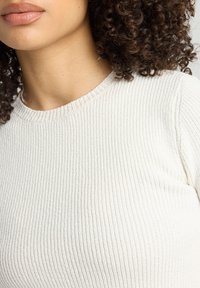 White ribbed knit sweater with a round neckline, featuring a textured surface and subtle stitching along the collar and seams.