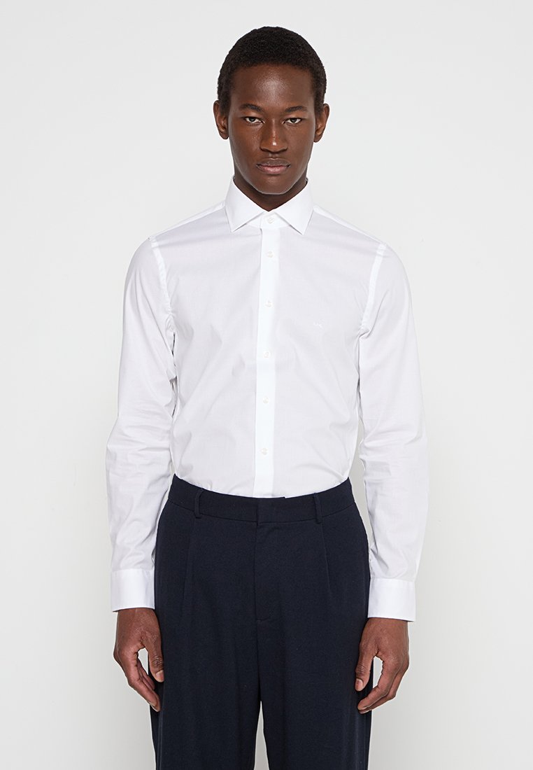 Camisa blanca de vestir con un diseño ajustado, frente abotonado, mangas largas y tela suave; combinada con pantalones oscuros, de pie frente a un fondo liso.