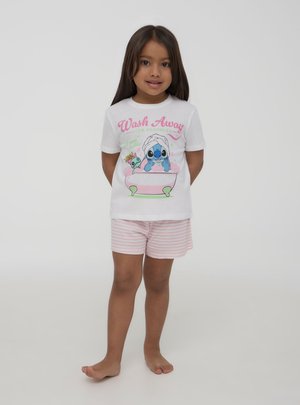 Ragazza giovane in piedi scalza che indossa una maglietta bianca con un personaggio dei cartoni animati nella vasca da bagno e pantaloncini rosa a righe, sorridendo alla fotocamera.