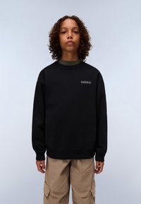 Sudadera negra de cuello redondo y puños de canalé. Presenta un pequeño logo en el pecho. Se lleva sobre una camiseta de manga larga verde y combina con pantalones beige.