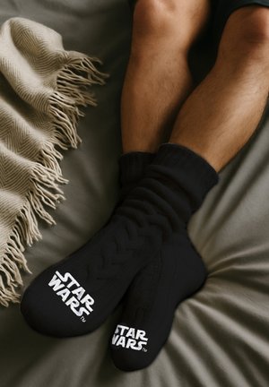 Schwarze gestrickte Socken mit einem strukturierten Design und dem weißen "STAR WARS"-Logo auf der Vorderseite, auf einer grauen Bettdecke neben einer Fransen-Decke liegend.
