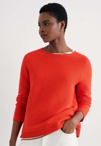 Maglione rosso a coste con colletto rotondo e maniche lunghe, caratterizzato da una vestibilità ampia e un tessuto testurizzato. Canottiera bianca visibile al colletto.