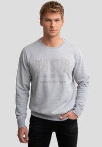Ung mand med kort blondt hår iført en lysegrå sweatshirt med teksten "BIG STAR You Life Your Jeans", stående foran en ensfarvet baggrund.