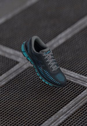 Schwarzer und blauer Asics Laufschuh mit strukturierter Sohle, der auf einer Metallgitteroberfläche mit sich kreuzenden Linien liegt.