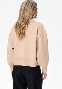 Beżowy sweter dzianinowy z opadającymi ramionami i prążkowanym wykończeniem. Posiada bufiaste rękawy i skrócony fason, co zapewnia luźniejszy krój. Widoczne czarne spodnie.