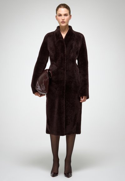 Garš, tumši sarkans mētelis ar mīkstu, teksturētu virsmu, platas piedurknes un apkakli, valkāts kopā ar tumšām zeķubiksēm un spīdīgām augstpapēžu kurpēm. Rokās tur saskaņotu somiņu.