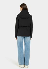 Musta lyhyt takki hupulla, jonka materiaalissa on tekstuuria ja säädettävä vyötärö, yhdistettynä vaaleansinisiin leveälahkeisiin farkkuihin ja valkoisiin tennareihin.