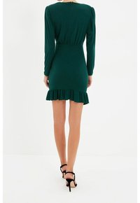 Femme portant une robe longue ajustée vert foncé à manches longues avec un ourlet à volants et des sandales noires à talons hauts, vue de dos.