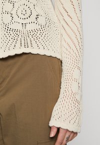 Tröja i krämfärg med ett intrikat öppet mönster, med långa ärmar och vågkant, tillsammans med bruna tygbyxor.