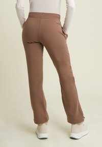 Persona vestita con pantaloni marroni aderenti e sneakers beige con suole bianche, in piedi con le mani in tasca contro uno sfondo chiaro e semplice.