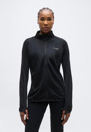 Ženska z pletenicami, oblečena v črno Columbia jakno z zadrgo in črne hlače, stoji pred belim ozadjem.