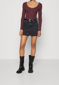 Kvinna som bär en vinröd långärmad topp, svart denim minikjol med ett bälte och svarta knähöga stövlar, stående mot en plain bakgrund.