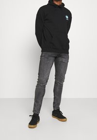 Černá mikina s kapucí a přední kapsou, zdobená modrou grafickou výzdobou na hrudi; spárována se šedými úzkými džíny a černými teniskami.