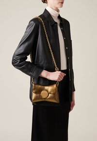 Metallic gold handbag with a chain strap, soft leather texture, and a circular accent on the front, worn over a black outfit.
