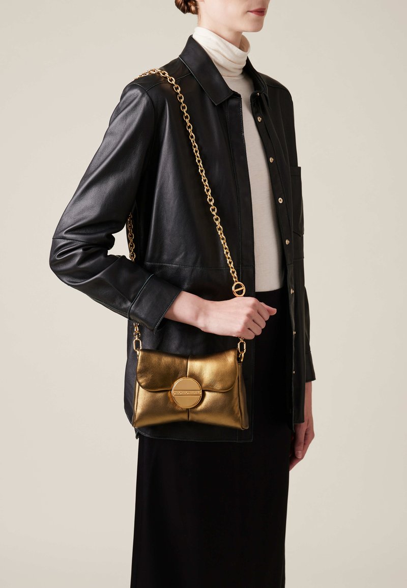 Metallic gold handbag with a chain strap, soft leather texture, and a circular accent on the front, worn over a black outfit.