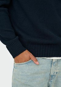 Maglione lavorato a maglia blu navy con polsini a coste, abbinato a jeans chiari strappati. Mano appoggiata nella tasca dei jeans.