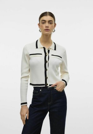 Cardigan bianco con strisce nere lungo il colletto e la patta anteriore, con bottoni dorati e polsini a coste fitted. Jeans in denim scuro.