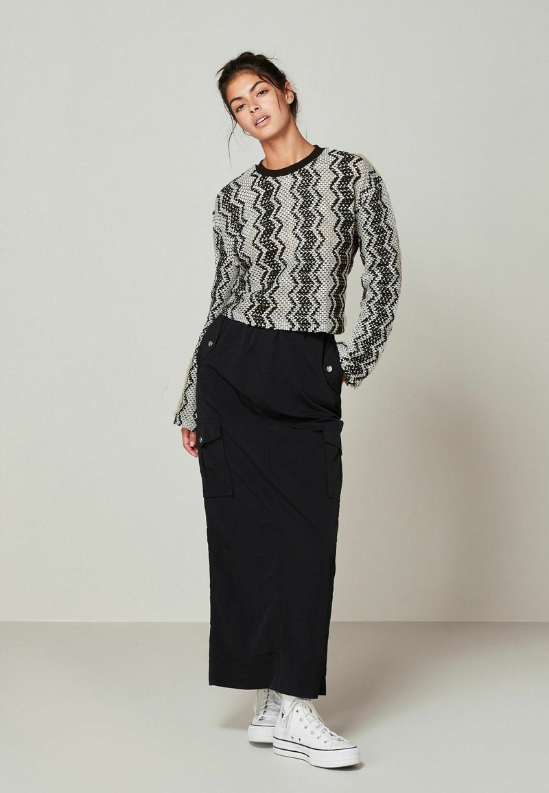 Černá maxi sukně s cargo kapsami, spárovaná s přiléhavým svetrem s černo-bílým zigzag vzorem. Celý outfit doplňují bílé šněrovací tenisky.