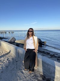 Beżowy długi płaszcz, biała koszula, czarne szerokie spodnie i brązowe sandały. Stoi na piaszczystej plaży obok kamiennego muru i wody. Słoneczny dzień.