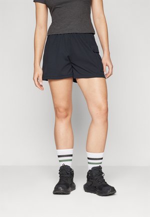 Žena oblečená v černých cargo šortkách, černých sportovních botách, bílých ponožkách se zelenými a černými pruhy a tmavě šedém topu s krátkými rukávy.