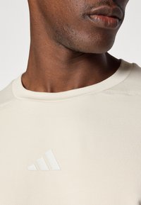 T-shirt de sport beige clair avec un col rond et le logo Adidas en ton sur ton blanc sur la poitrine. Texture lisse avec un tissu extensible.