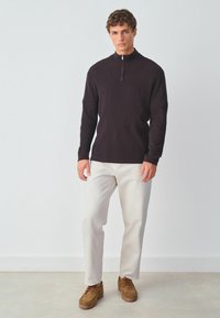 Donker gebreide pullover met een hoge kraag en een kwart-rits, voorzien van een gestructureerd ruitpatroon. Gekleed met lichtgekleurde broeken en bruine schoenen.