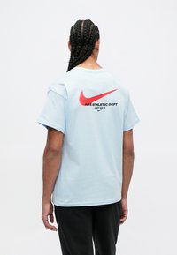 Cilvēks ar garām pītām matēm, tērpies gaiši zilos T-kreklos ar sarkano Nike swoosh un melniem burtiem, kas uzrakstīti aizmugurē: "Nike Athletic Dept Just Do It".