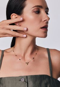 Złoty naszyjnik z czarnymi koralikami, z trójkątnym wisiorkiem. Modelka ma na sobie zielony top; widoczna ozdobna kolczyk w uchu. Gładka tekstura.