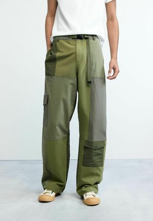 Mees, kes kannab rohelise toonides lahtisi tikkimisest koostatud lahtiseid pükse suurte taskutega, ühendatuna valgete tossude ja valge särgiga.