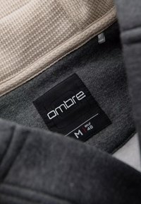 Grå sweatshirt indvendig med et struktureret beige stof ved halsen. Har et sort mærke med "ombre" og størrelsesoplysninger.