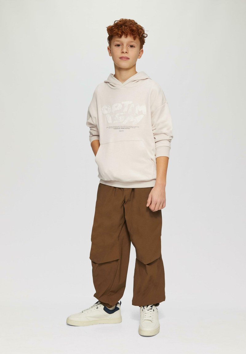 Lichtbeige hoodie met de opdruk "OPTIMISME", gecombineerd met losse bruine cargobroek en witte sneakers met marineblauwe accenten. Gladde stof, relaxte pasvorm.