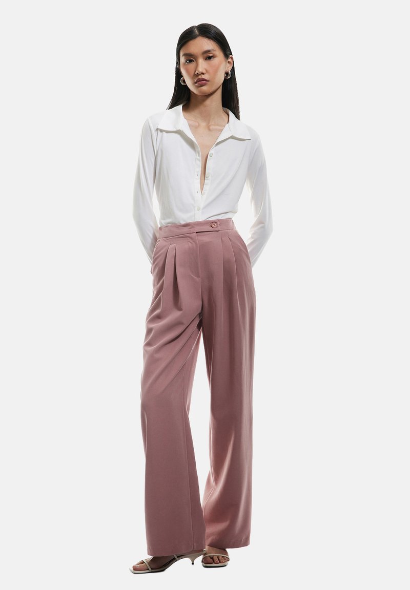 Valkoinen pitkähihaisten pusero, jossa on nappisolki, yhdistetty korkeavyötäröisiin, laskostettuihin vaaleanpunaiseen housuun ja vaaleisiin korkokenkiin.