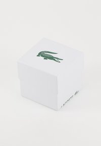 Caixa quadrada branca com superfície texturizada. Logotipo de crocodilo verde em cima e nas laterais. Design simples e limpo, sem padrões ou detalhes adicionais.