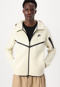 Veste à capuche Nike en tissu blanc cassé avec des accents noirs. Caractéristiques : col haut, fermeture éclair et poches latérales. Texture lisse.
