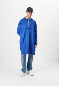 Blå huvjacka med framficka, elastisk snörning vid halsen och randig design på bröstet, kombinerad med ljusa jeans och vita skor.