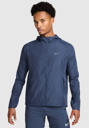 Marineblå Nike-jakke med hætte, lynlås foran og reflekterende logo. Har lange ærmer og et let, tekstureret stof.