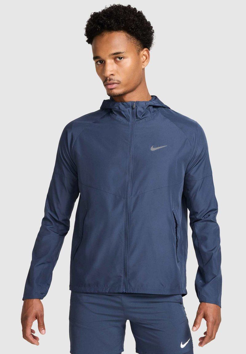 Granatowa kurtka Nike z kapturem, zapinana na zamek, z odblaskowym logo. Posiada długie rękawy i lekką, teksturowaną tkaninę.