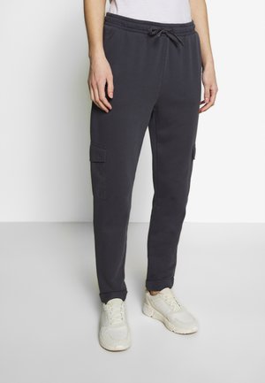 Personne portant un pantalon jogger cargo gris foncé avec des poches latérales et des baskets blanches, se tenant devant un fond clair uni.