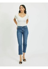 T-shirt branco em V, de manga curta, combinado com jeans azuis de cintura média e pernas retas, e sandálias pretas de salto aberto; design simples e casual.