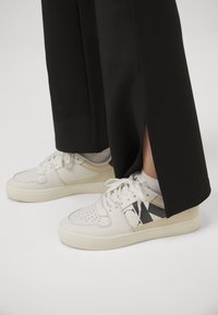 Vita lädersneakers med perforeringar, en tryckt logotyp i svart och platta skosnören. Bärs med svarta byxor med sprund vid fållen.