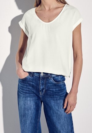 Femme portant un chemisier blanc uni à manches courtes rentré dans un jean bleu taille mi-haute, avec la main gauche dans la poche.