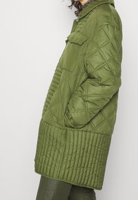 Chaqueta acolchada de color verde oliva con un alto cuello, bolsillos laterales y un diseño texturizado que presenta patrones de costura diagonales y horizontales.