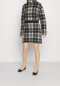 Ullblandad outfit med rutmönster i marinblått och krämfärgat, hög krage och långa ärmar med figursydd midja och matchande kjol. Svarta ballerinaskor.