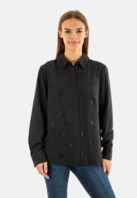 Camicia nera con bottoni e maniche lunghe, caratterizzata da un sottile ricamo floreale nero e una texture liscia. Indossata con jeans in denim blu.